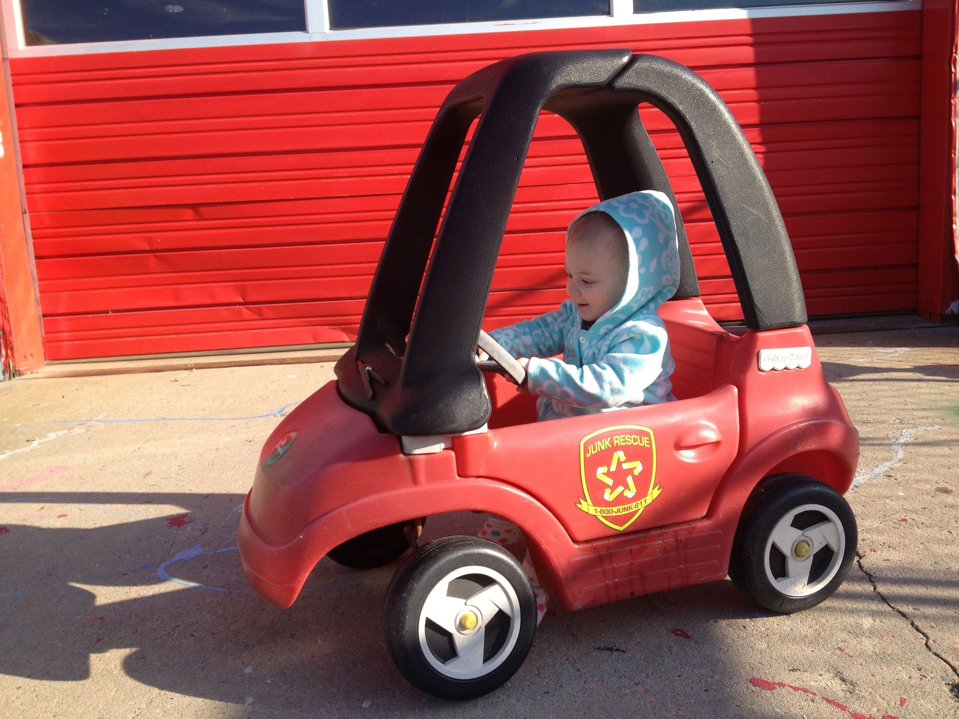 Child in blue hoodie driving a red Little Tikes car with a Fire Patrol emblem.