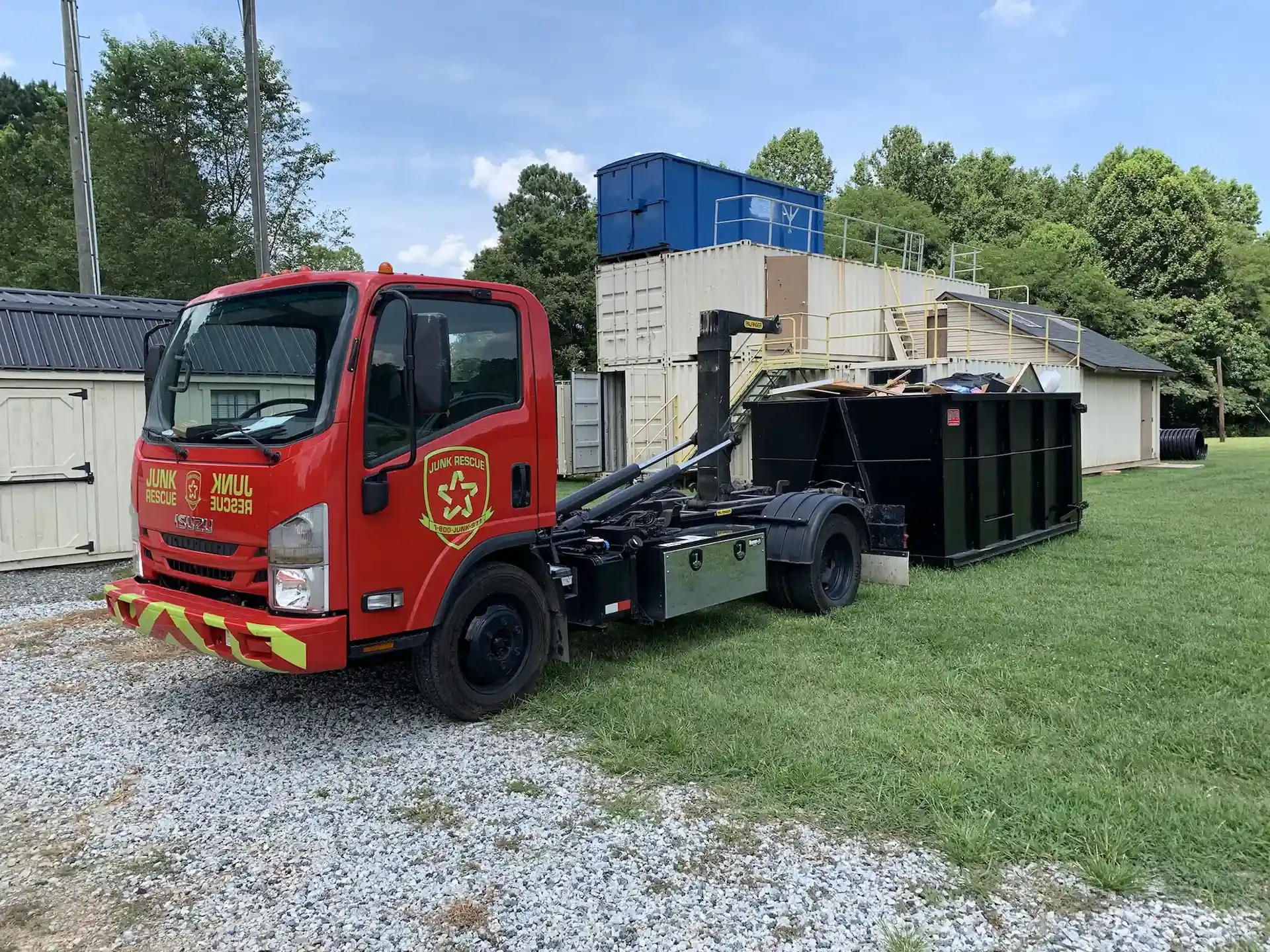 Red truck with a black dumpster on a grassy area near buildings.