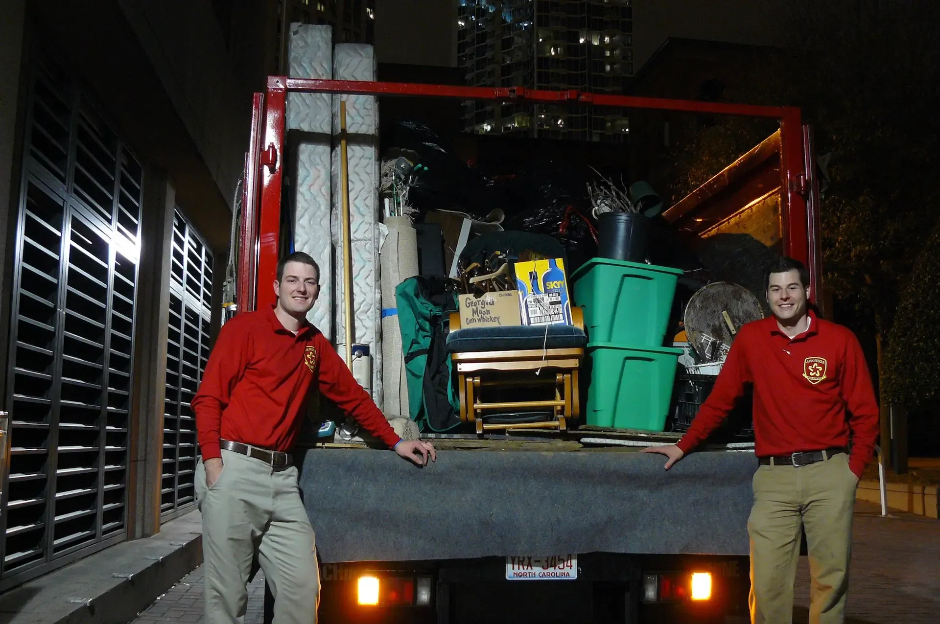 Two men in red shirts and khaki pants stand in front of a moving truck at night, loaded with items.