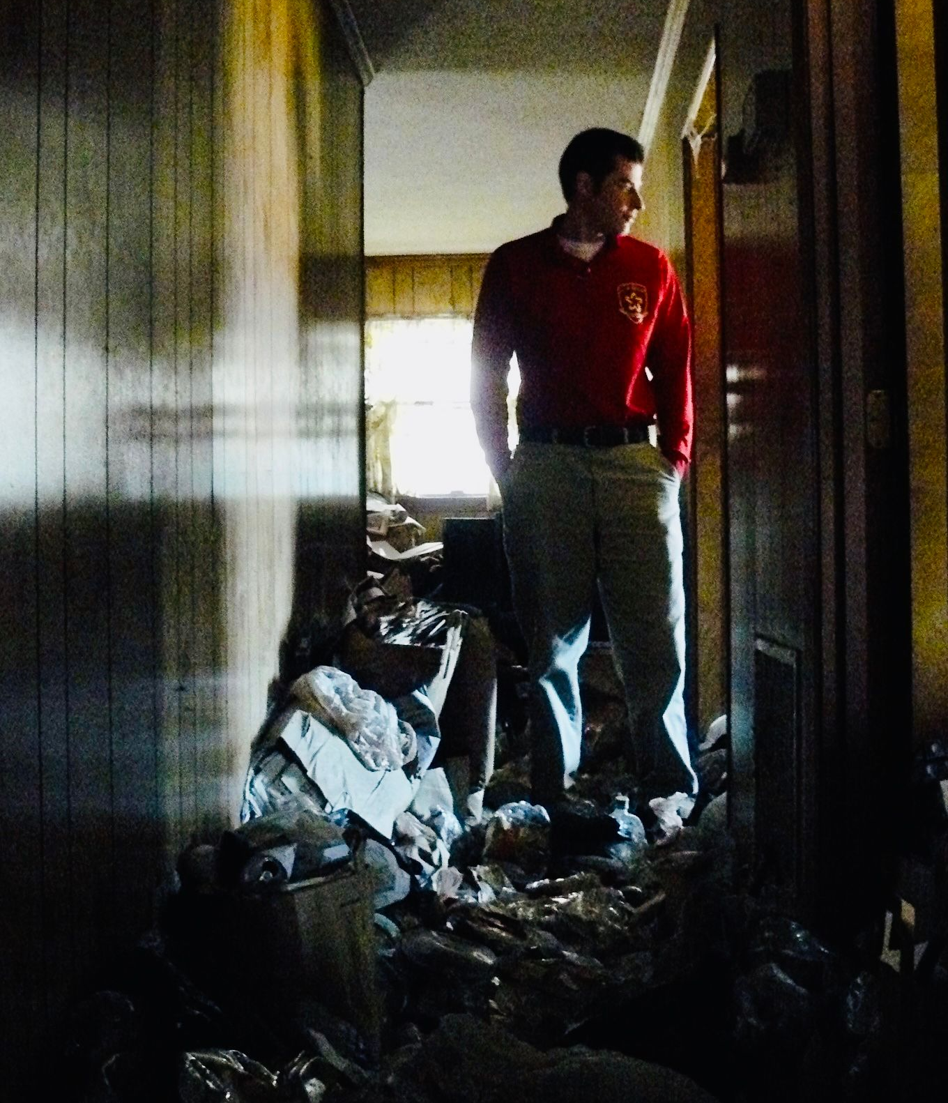 Man in red shirt stands in debris-filled hallway, looking towards bright light.