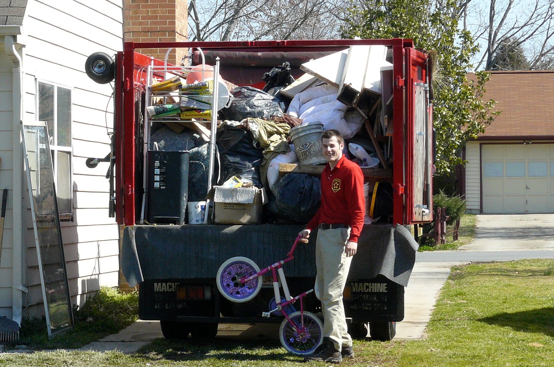 Man stands beside a packed garbage truck. The truck bed is full of items; he holds a small, pink bicycle.