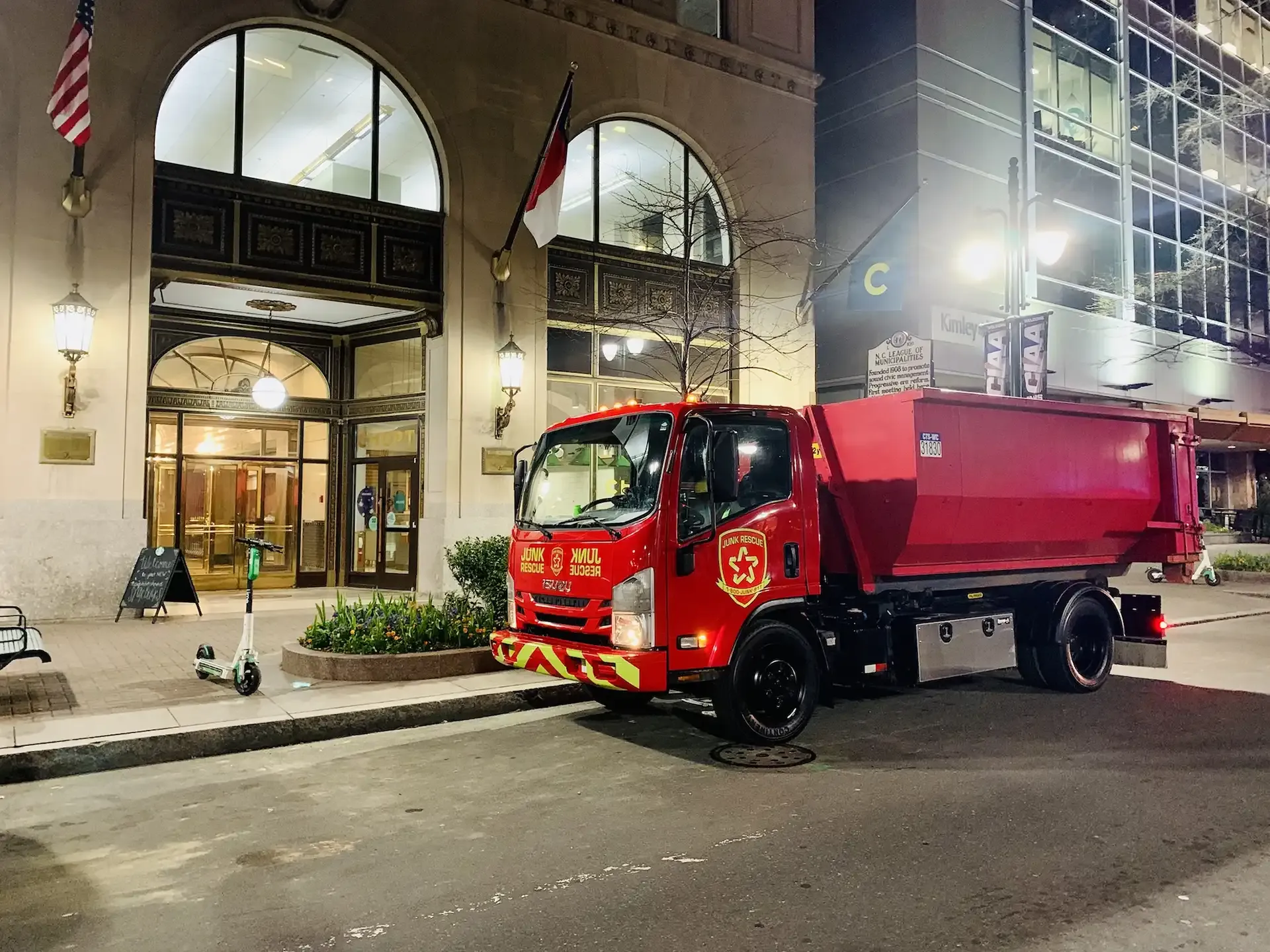 Red garbage truck parked outside a building with arched entrance and flags at night.