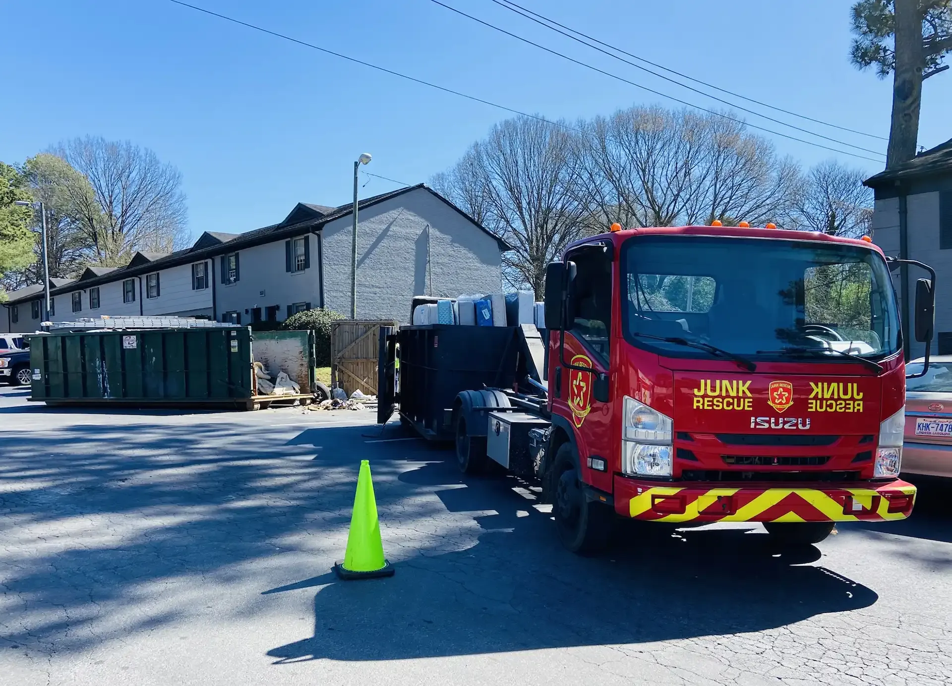 Red junk removal truck parked on a street near a green dumpster. A traffic cone is in front.