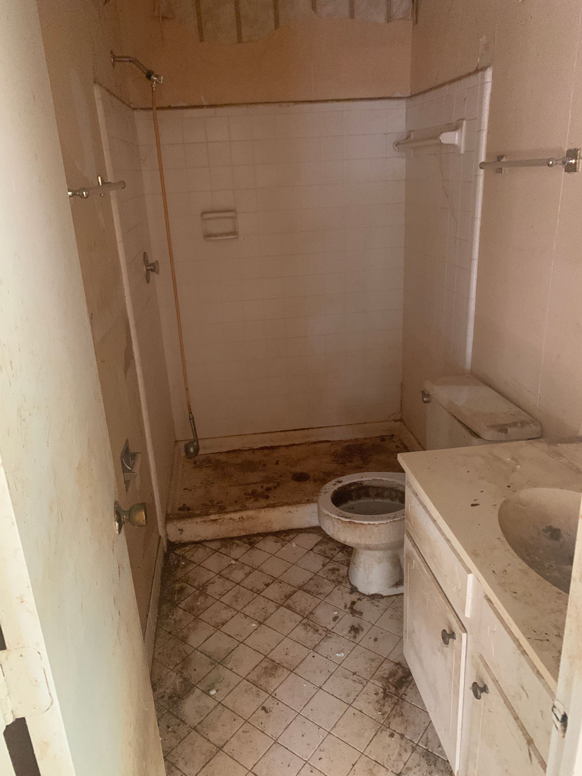 Dirty, old bathroom with a toilet, sink, and shower. Floor and walls are stained.