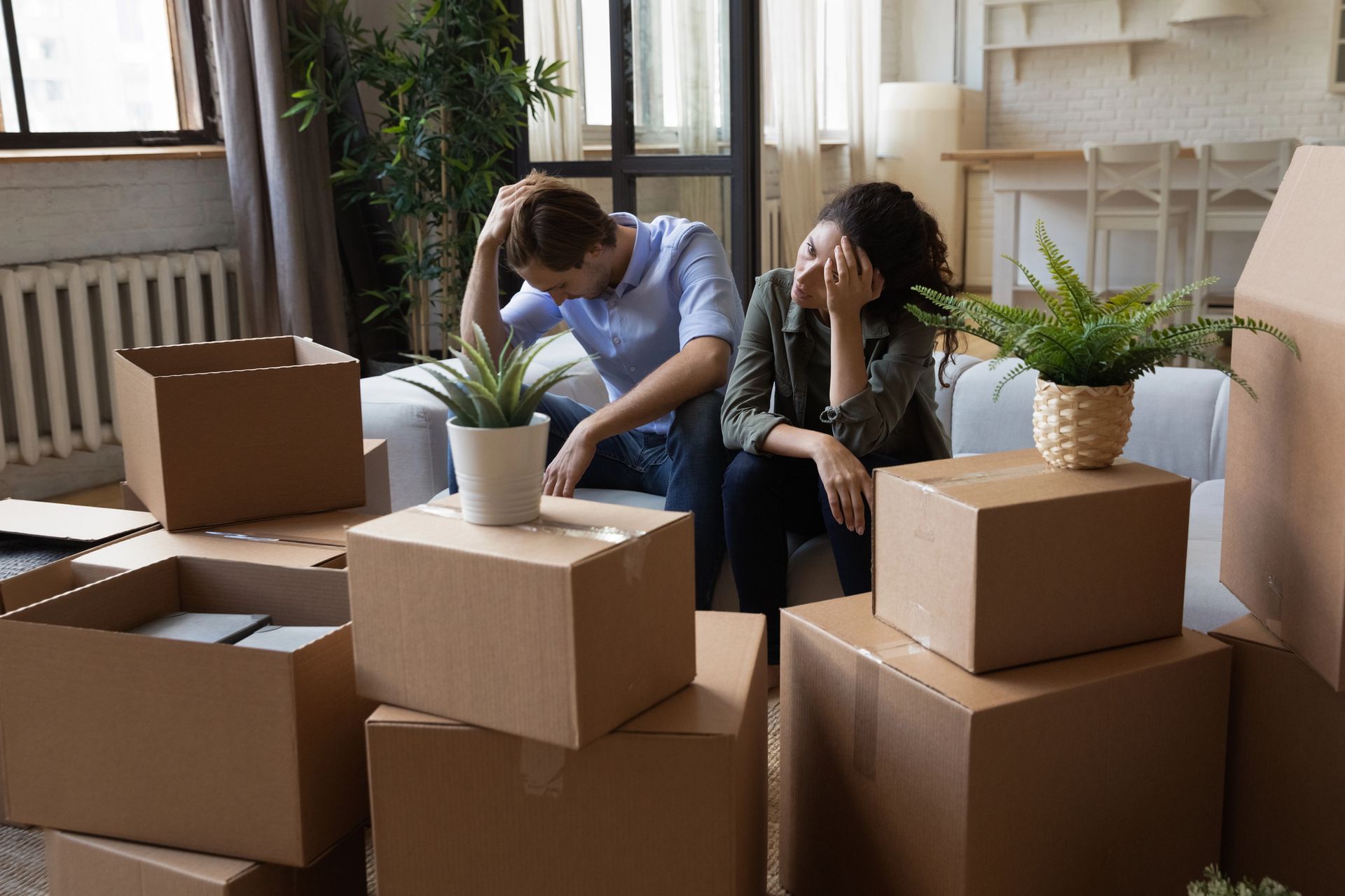 Two people sitting among stacked moving boxes in a bright room, looking tired during unpacking