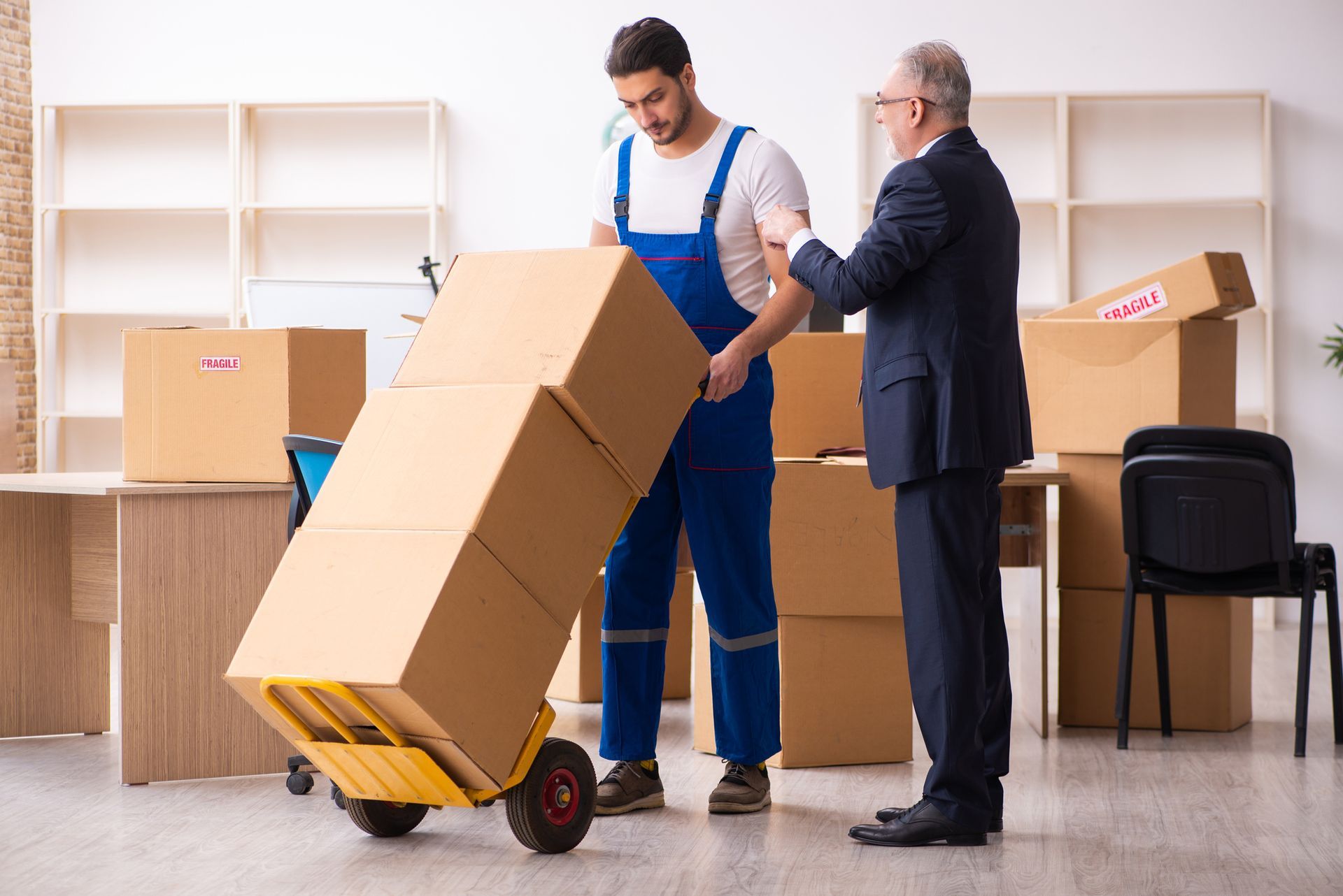 Two men moving a cardboard box on a dolly in a room with shelves and moving boxes.