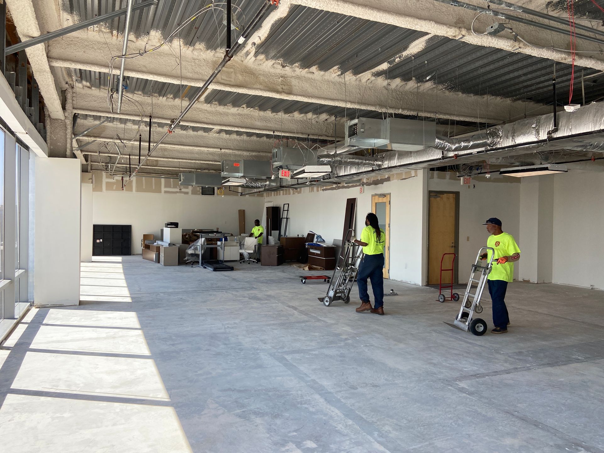 Workers in a large, unfinished room. One uses a hand truck, others stand. Concrete ceiling, bare floor, and natural light.
