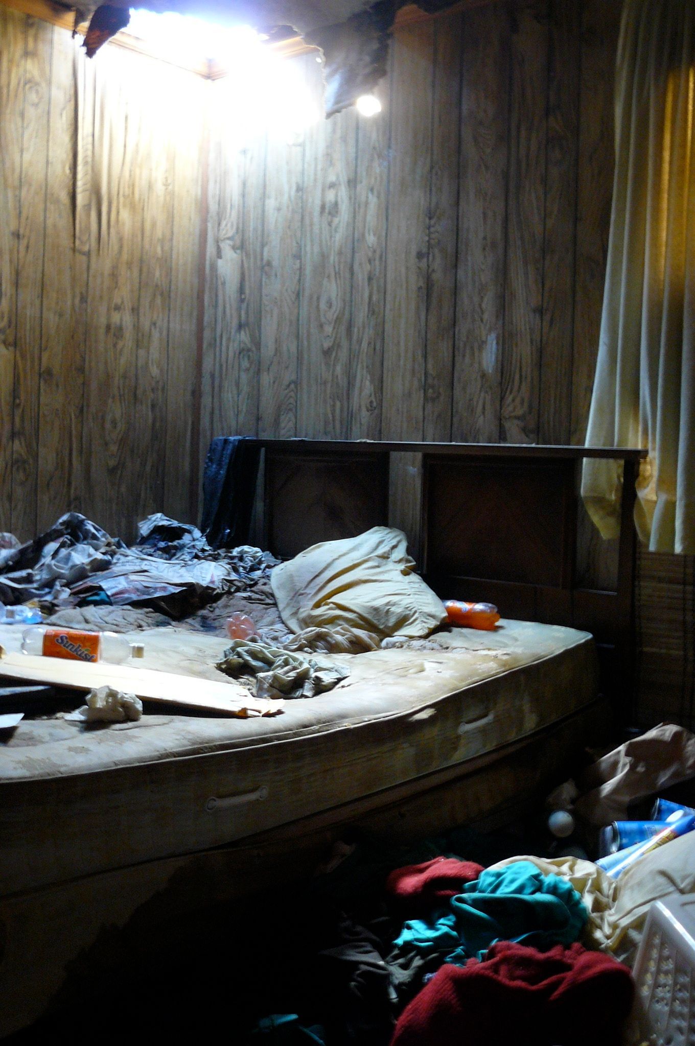 Brad Redden Messy room with a damaged ceiling, cluttered mattress, and piles of clothes.