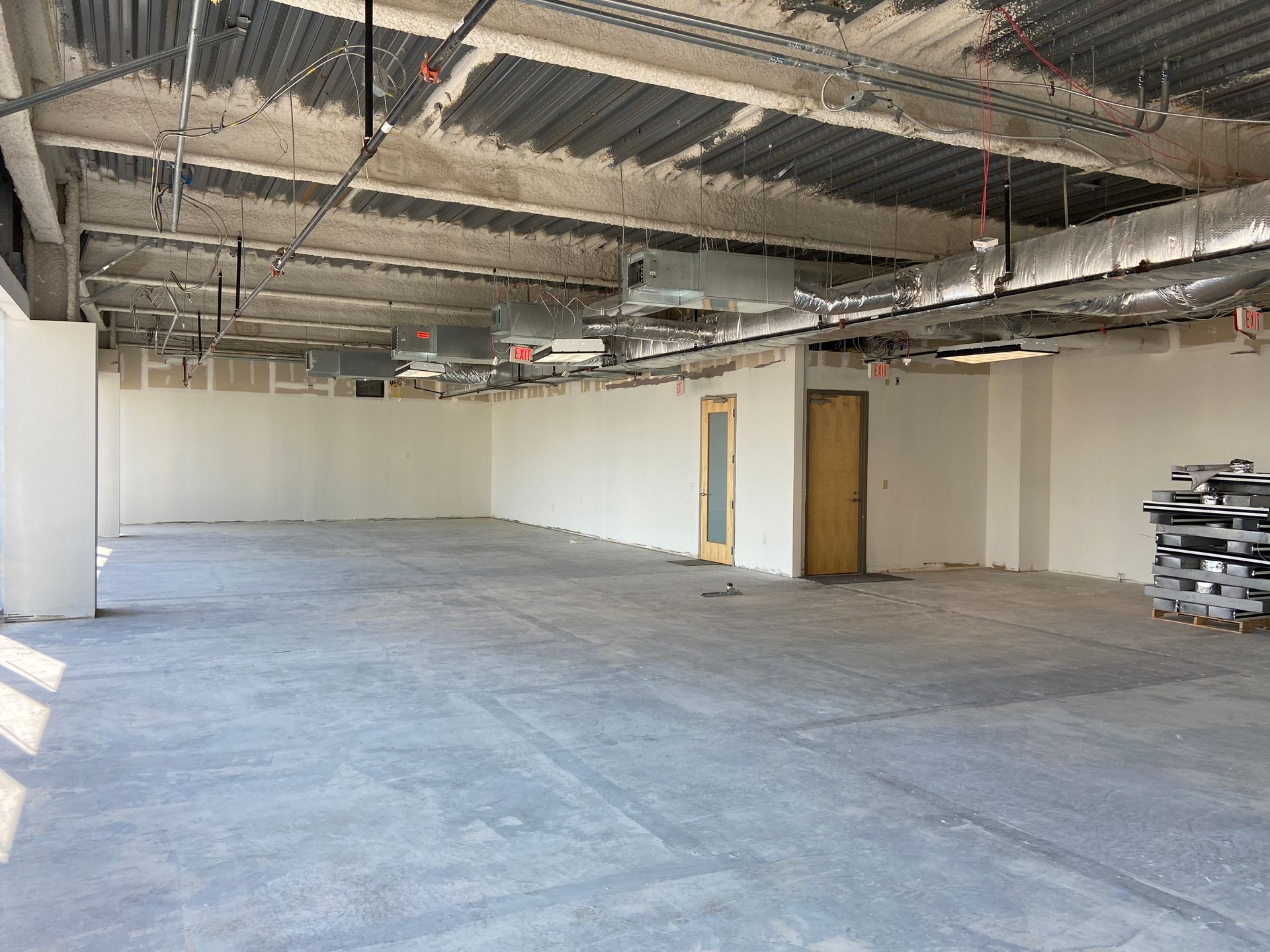 Interior of a commercial space under construction; gray concrete floor, white walls, exposed ceiling infrastructure.