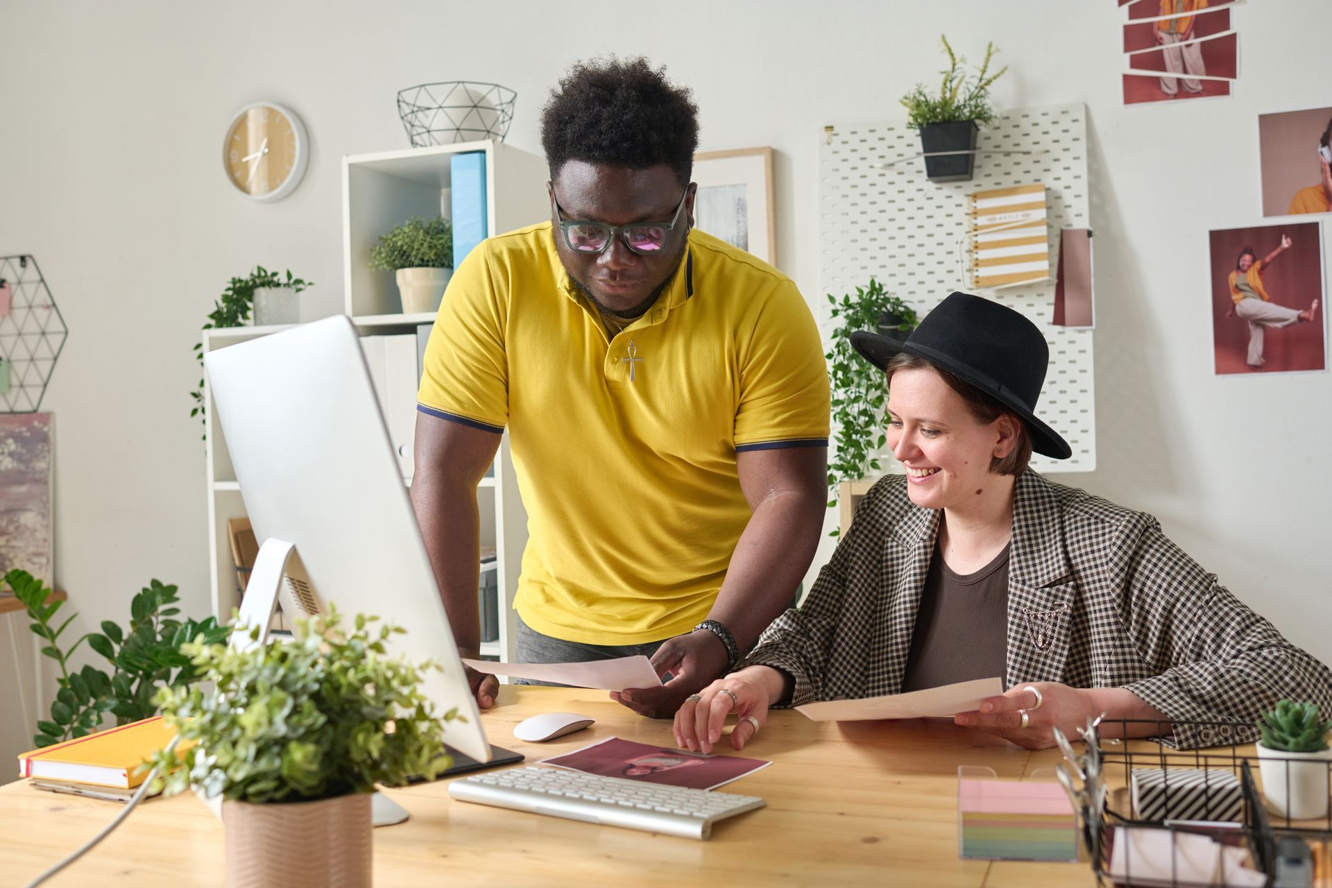 Two people reviewing papers at a desk, with a computer. One person wears a yellow shirt, the other a black hat.