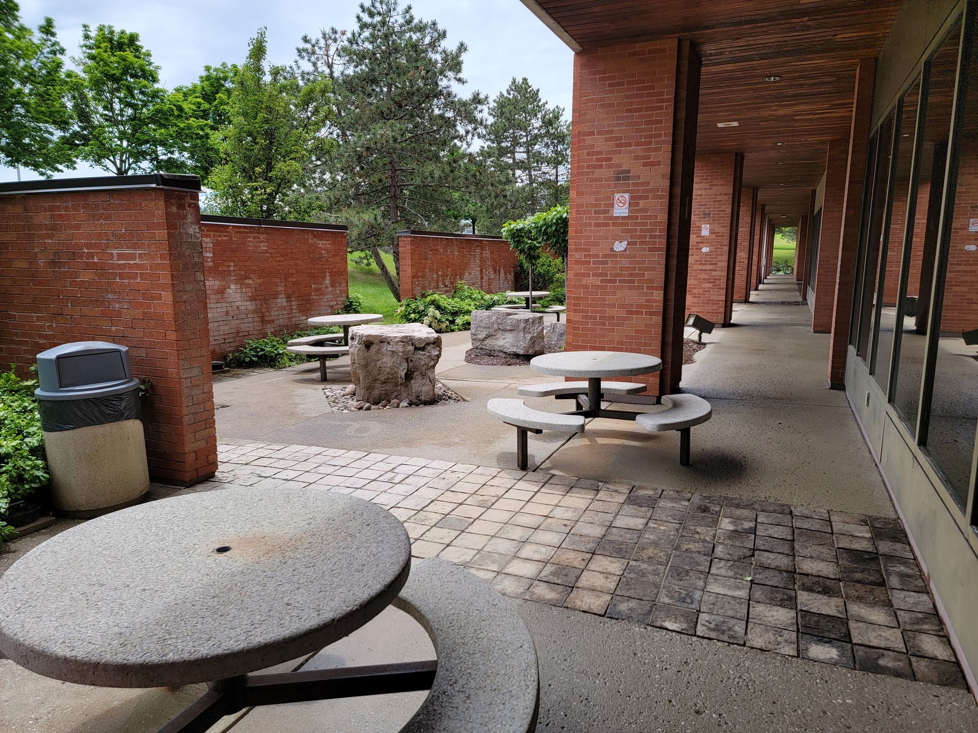 Outdoor brick patio with round concrete tables, waste bin, and walkway alongside a building with large windows.