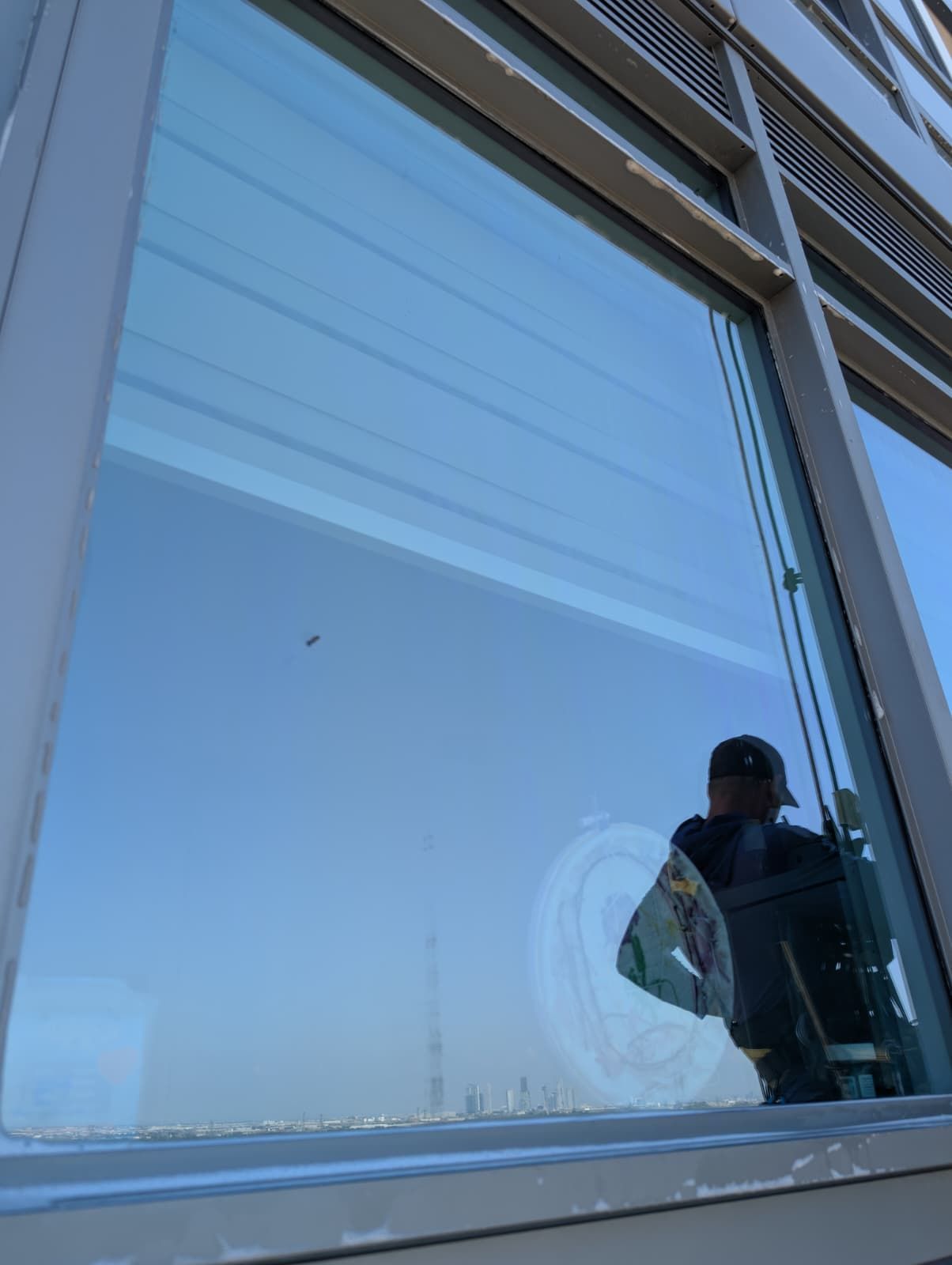 Person cleaning a large window on a tall building; reflection shows sky, city, and cleaning supplies.