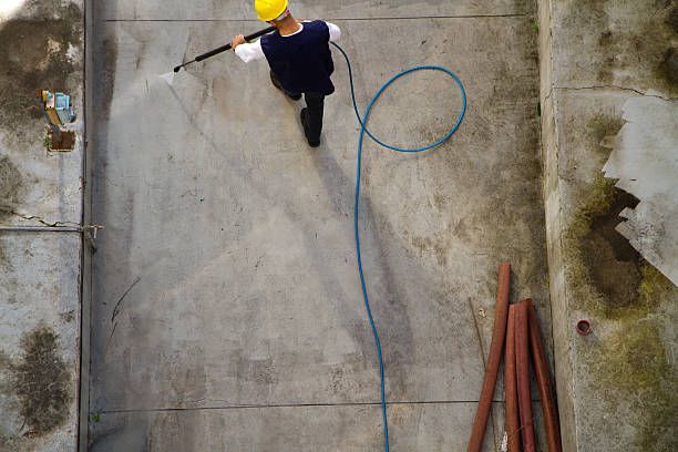 Worker in yellow hard hat power washes concrete floor.