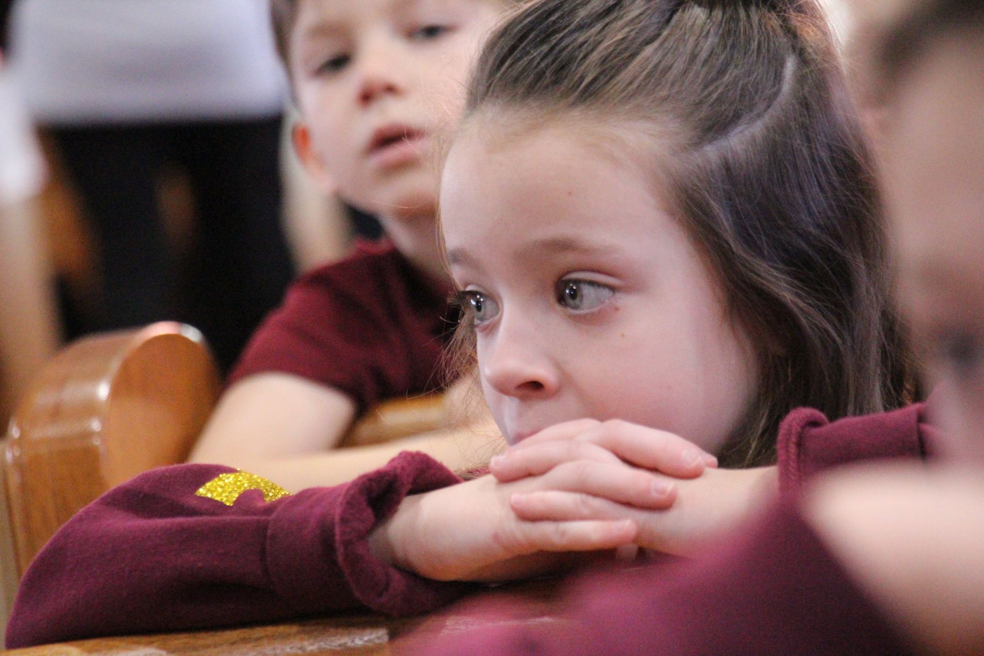 Child resting chin on folded hands in a classroom, watching intently; another child sits blurred behind.