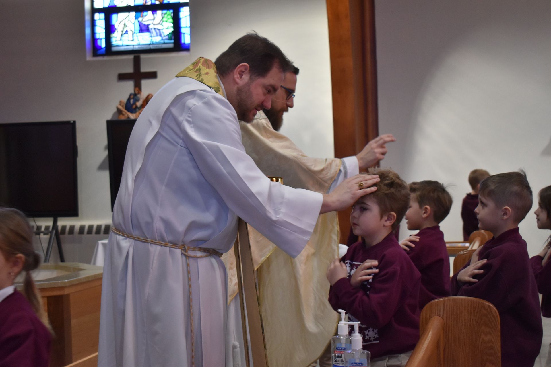 Priest blessing children in a church, with hands raised over a child in maroon uniform.