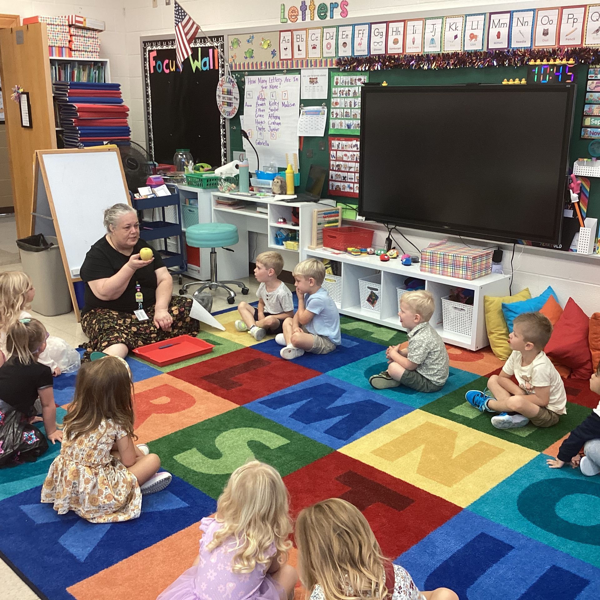 Teacher sitting on a colorful classroom rug, leading a small group of children in a circle activity.