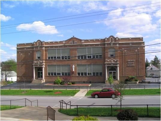 Brick school building with large windows and a red car parked in front under a blue sky.