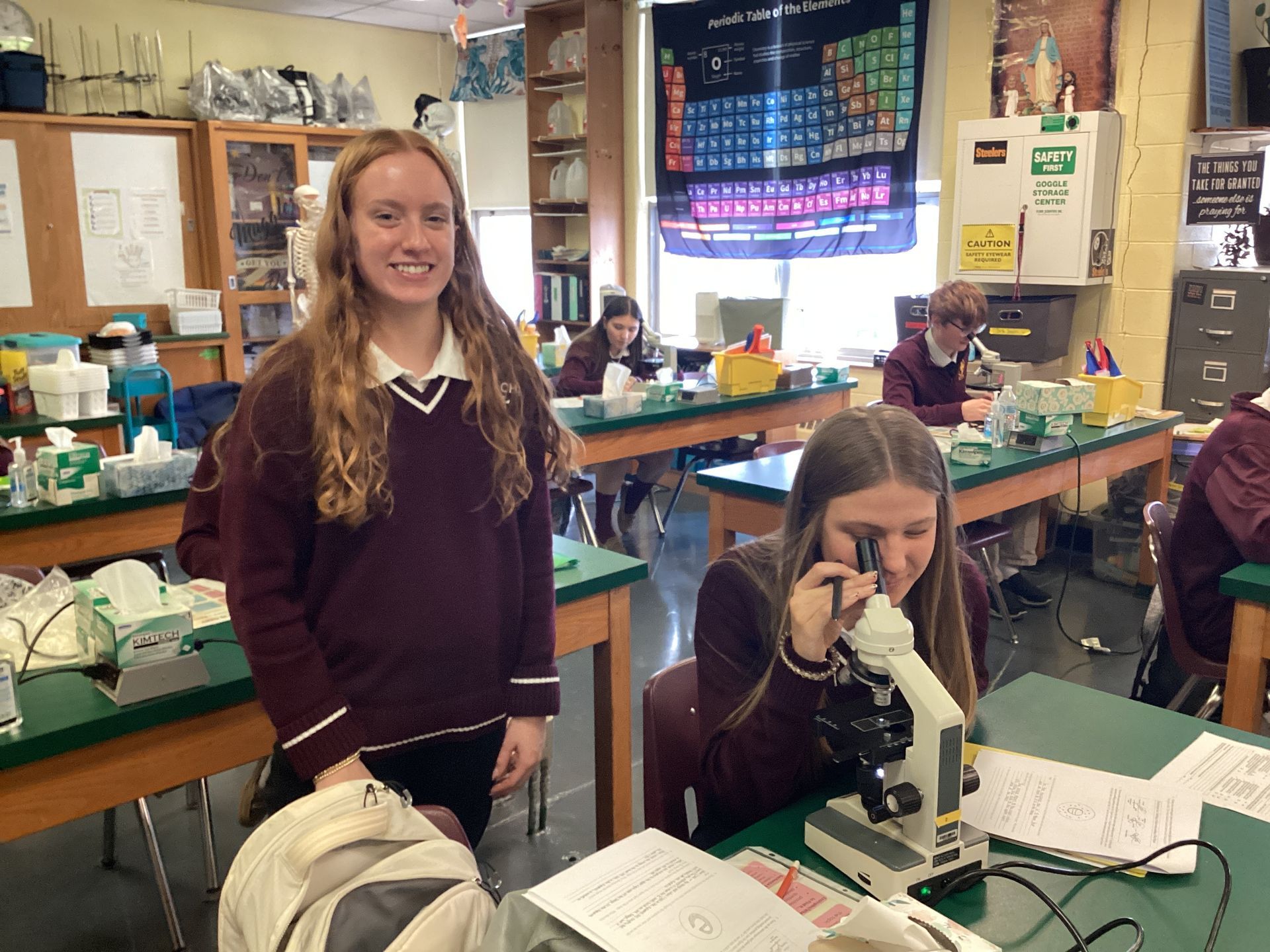 Two students in a classroom; one stands smiling while another looks through a microscope.