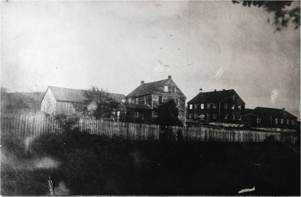 Black-and-white photo of a hillside with several old buildings behind a fence and open field