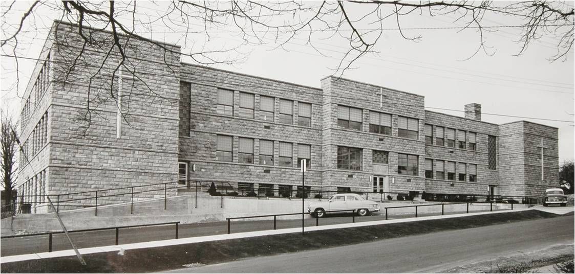 Black-and-white photo of a large brick building beside a snowy street with bare trees and parked cars.