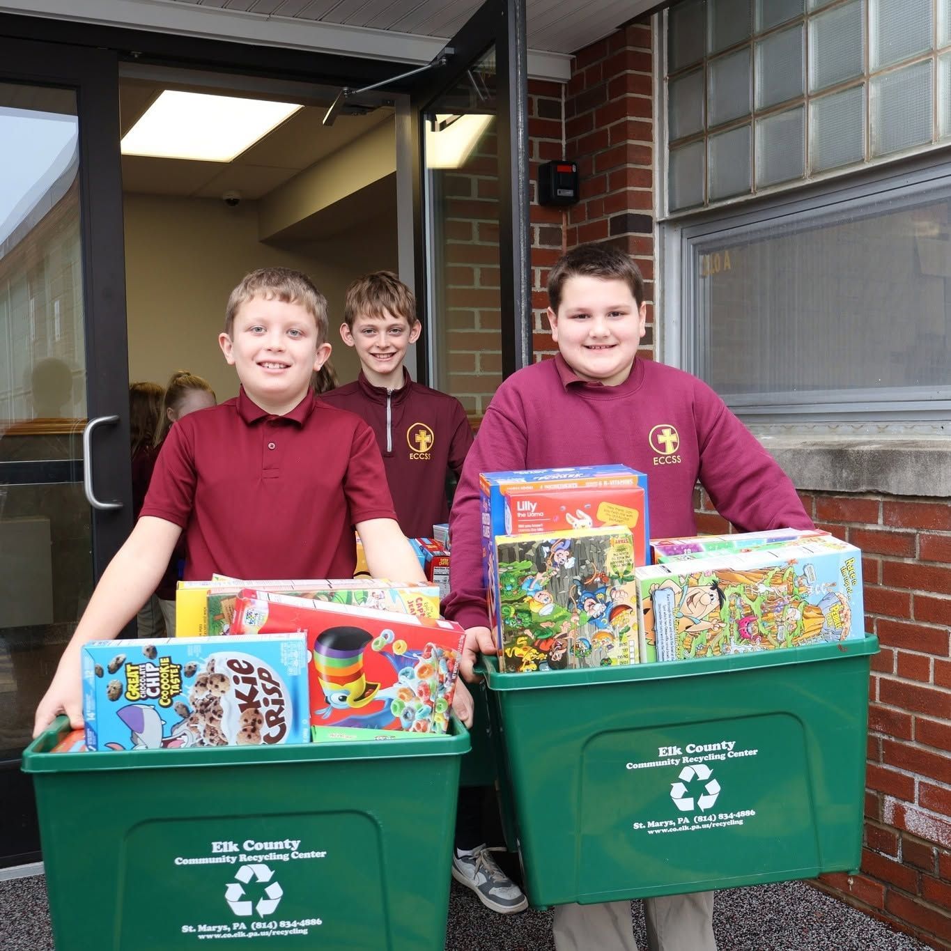 Three students in maroon shirts pushing bins of toys and books outside a building