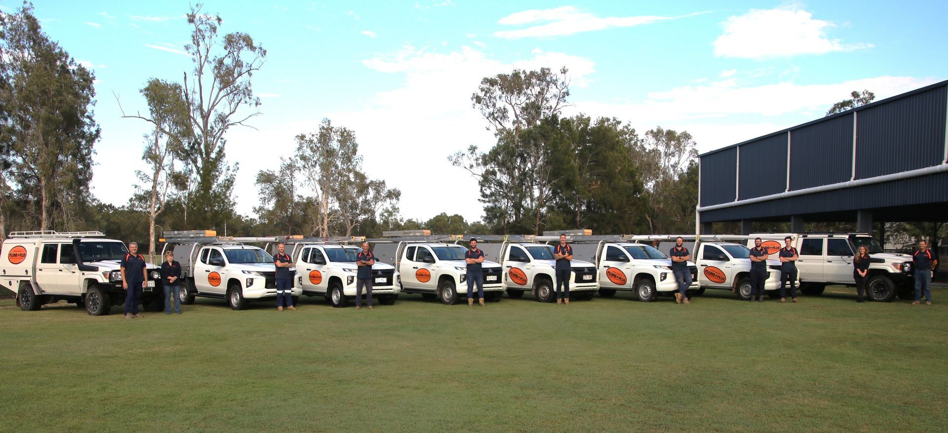 Row of white utility vehicles parked on a grassy field beside a building under a cloudy sky — PlumbPlus in Kawana, QLD