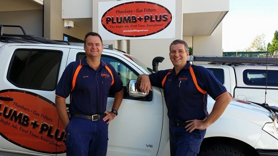 Two professional plumbers in navy and orange uniforms stand smiling in front of a service vehicle with Plumb+Plus branding.