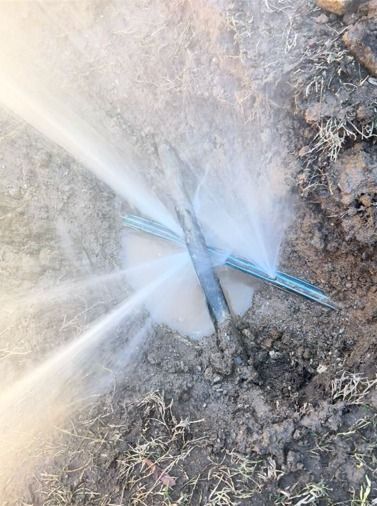 A high-pressure water leak spraying from a burst blue plastic pipe buried in dirt.