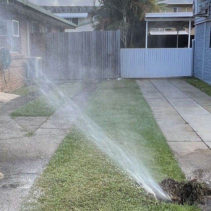 A residential lawn sprinkler spraying water across a strip of grass between a house and a paved driveway.