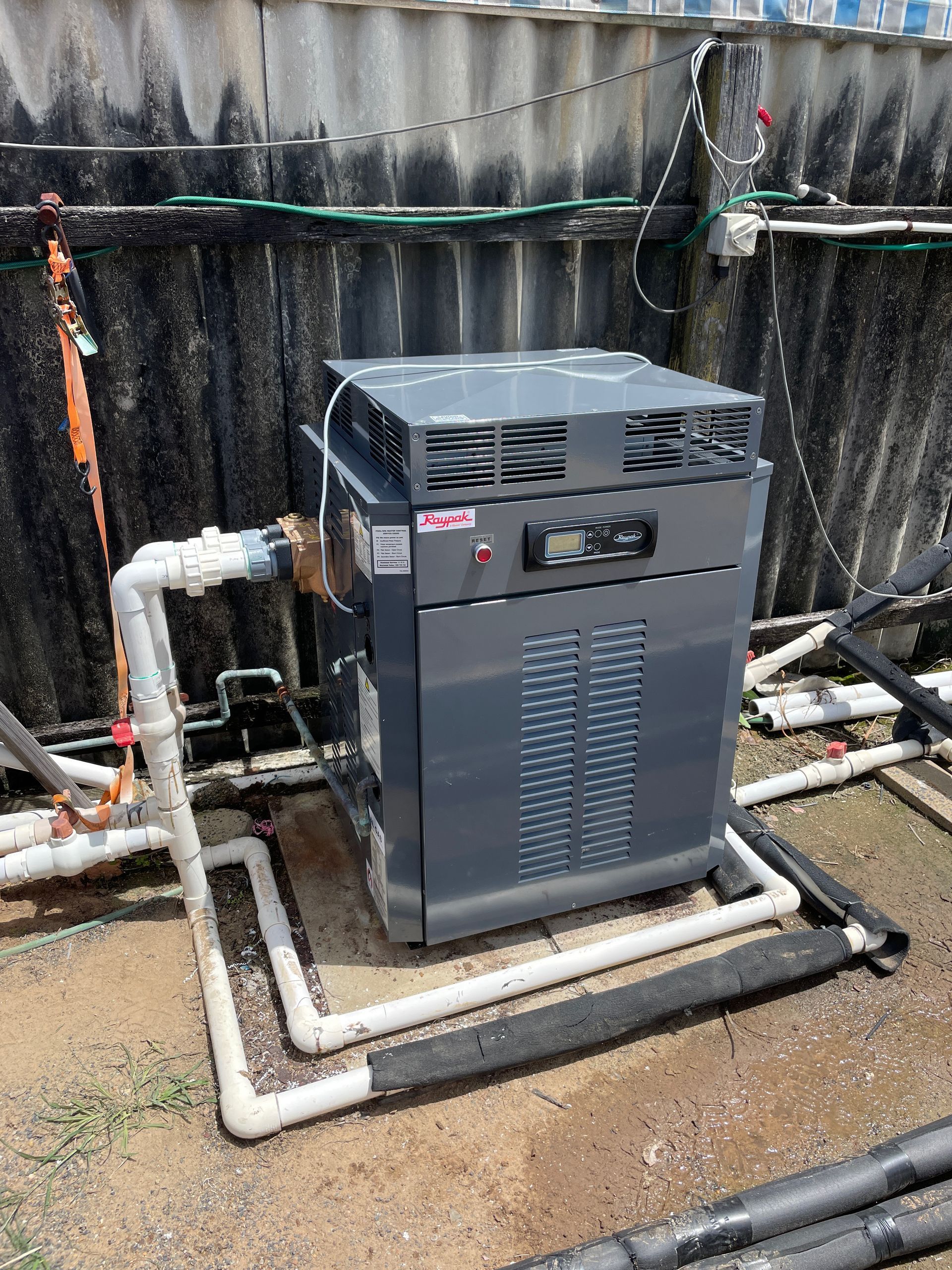 Grey outdoor HVAC unit with white pipes and hoses, installed beside a black fence on gravel.