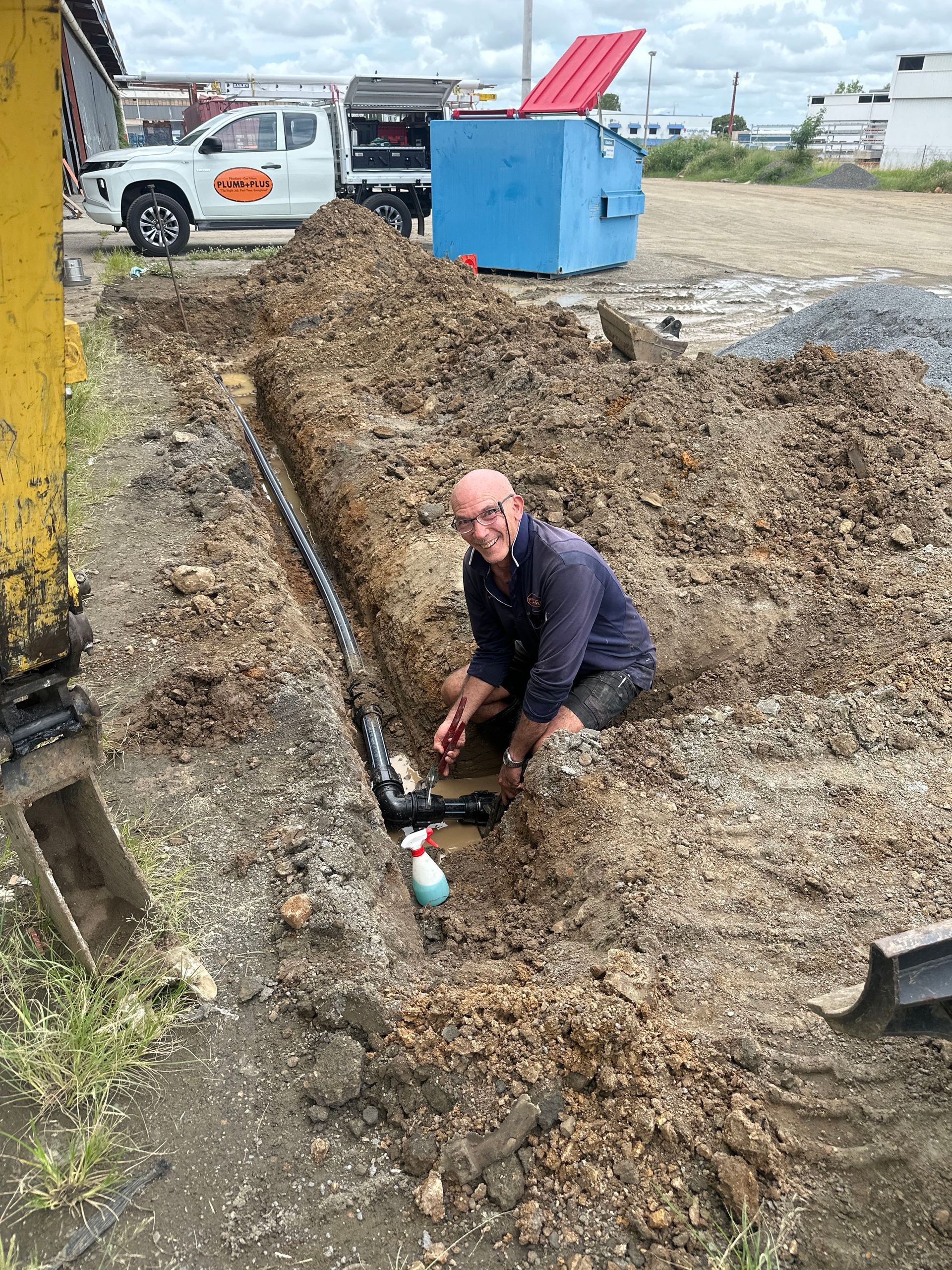 Worker crouches in a muddy trench beside a blue utility box on a construction site.