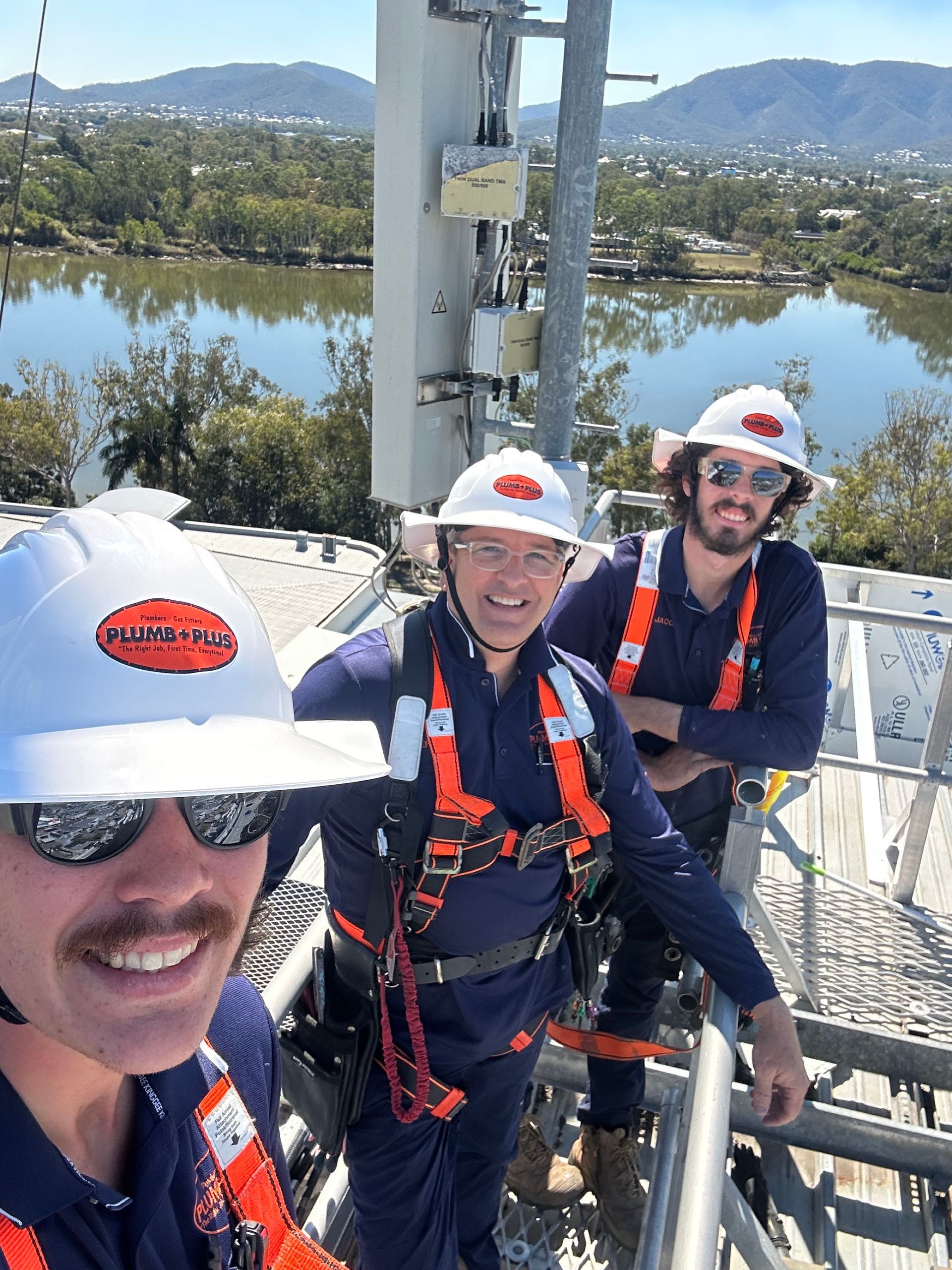 Three climbers in helmets and harnesses smiling on a metal tower with a river and hills behind them