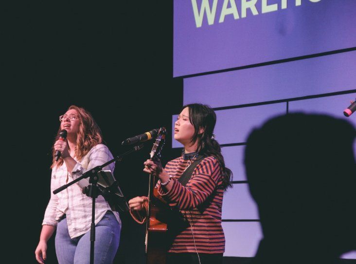 Two women singing into microphones on a stage in front of a sign that says warley