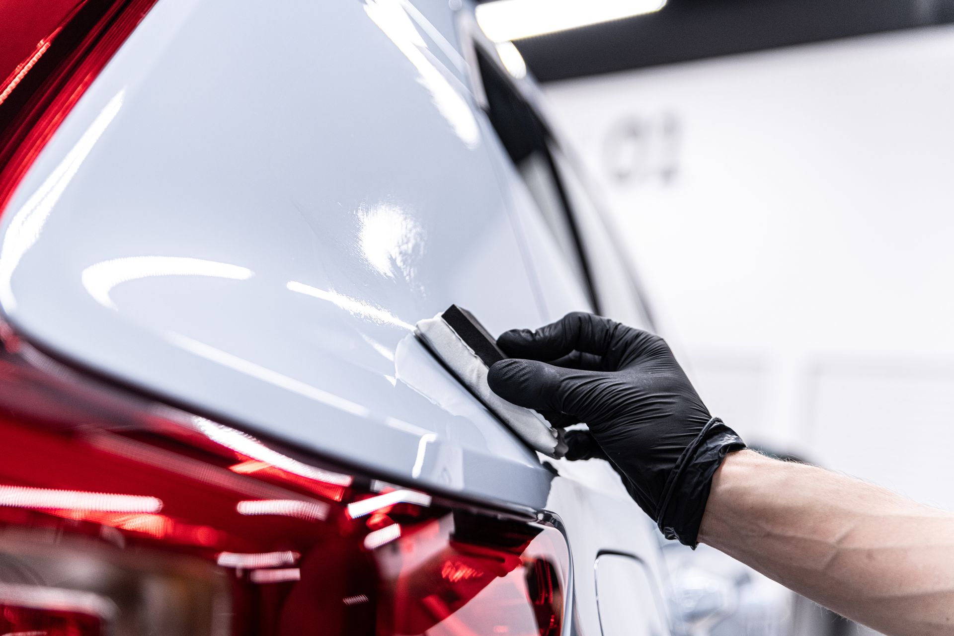 Gloved hand applying film to a white car’s rear panel in an auto detailing shop