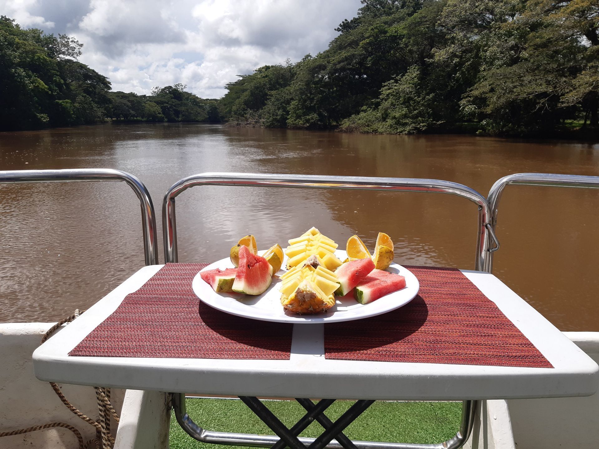Fruit plate on a table aboard a boat on a river, surrounded by trees.