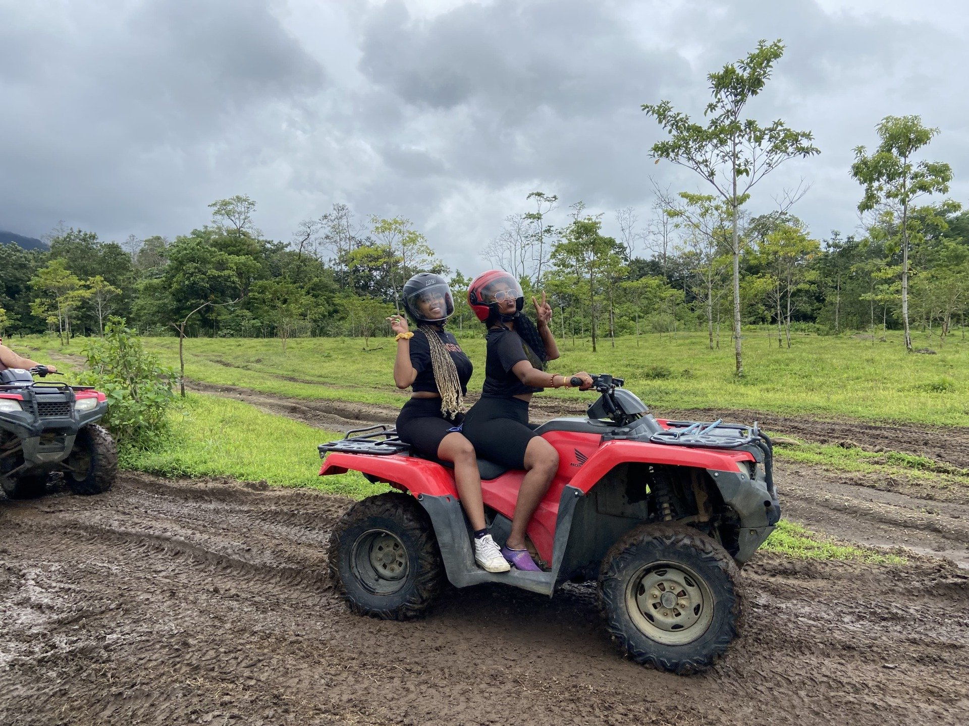 Two women on a red ATV, gesturing, on a muddy path, surrounded by green fields and trees.