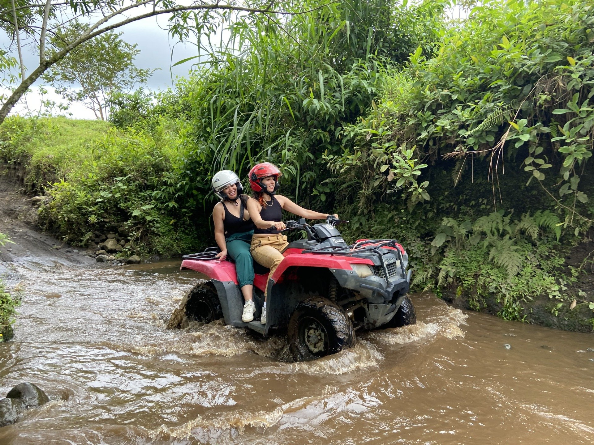Two women ride a red ATV through muddy water. They wear helmets and are in a lush, green setting.