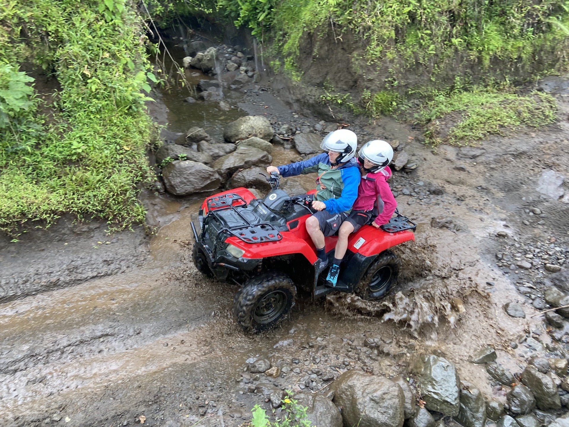 Two children on a red ATV driving through mud on a trail, both wearing helmets.