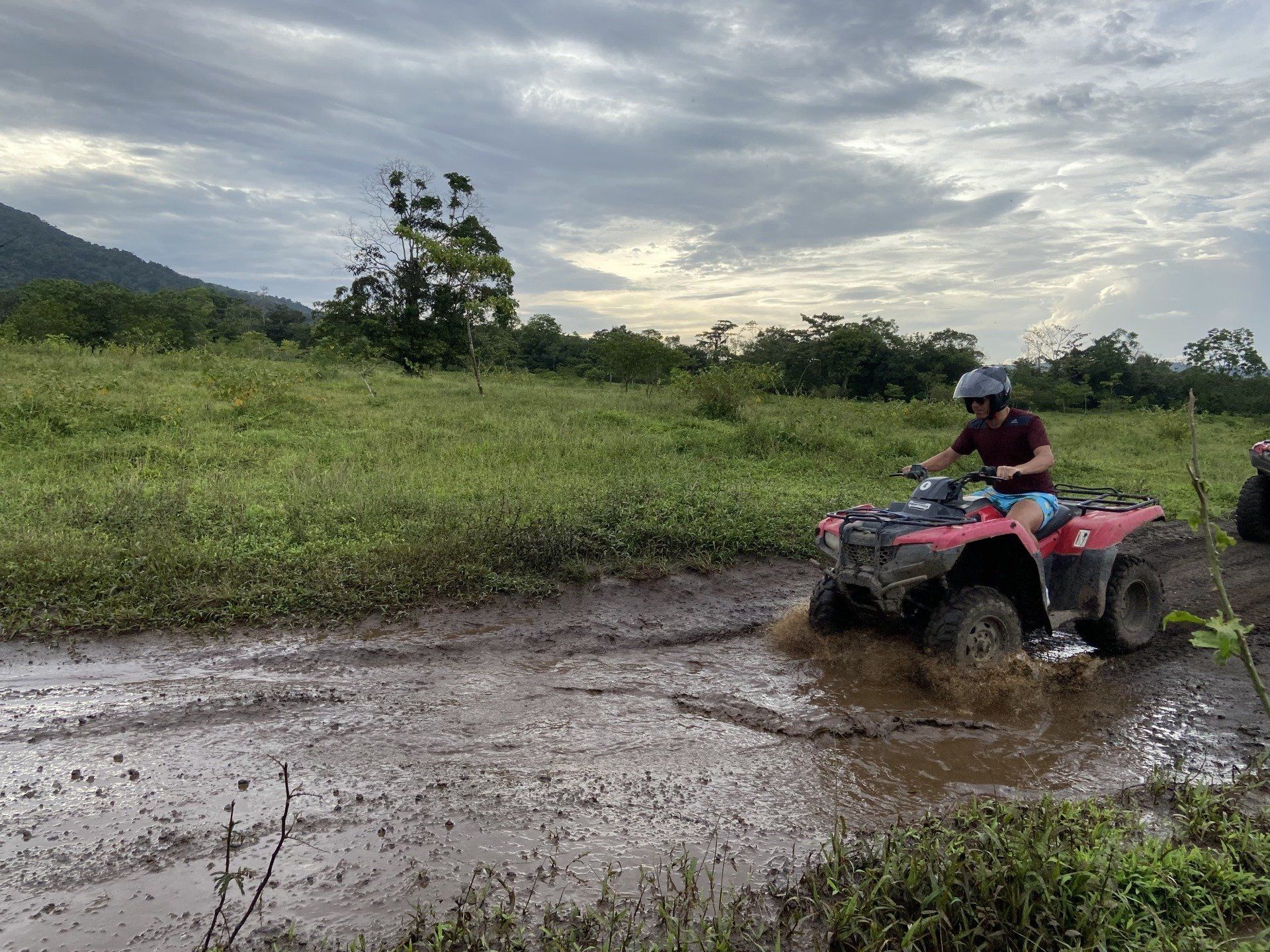 Person riding a red ATV through a muddy field under a cloudy sky.