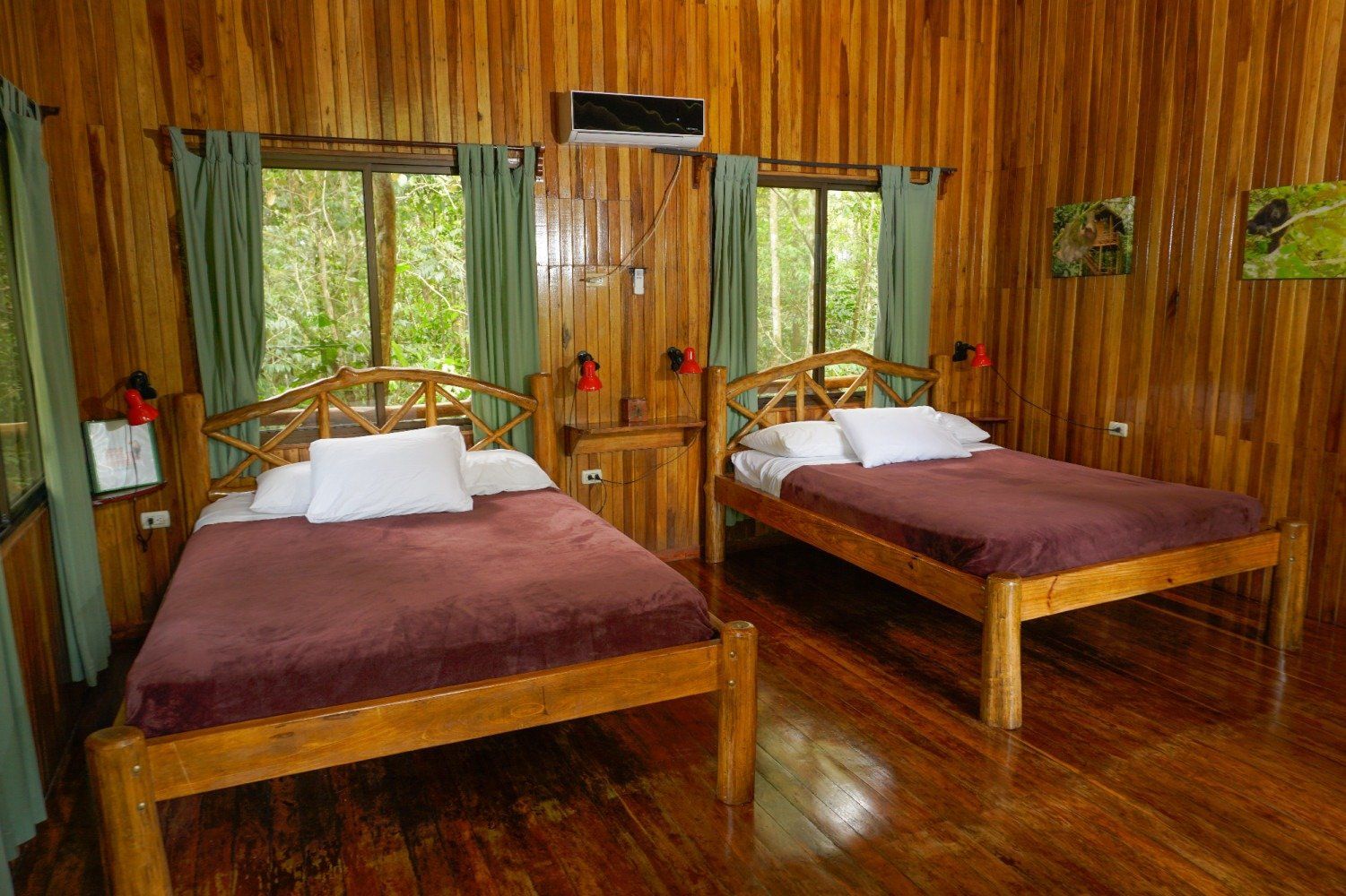 A wooden cabin bedroom with two beds, green curtains, and a forest view.