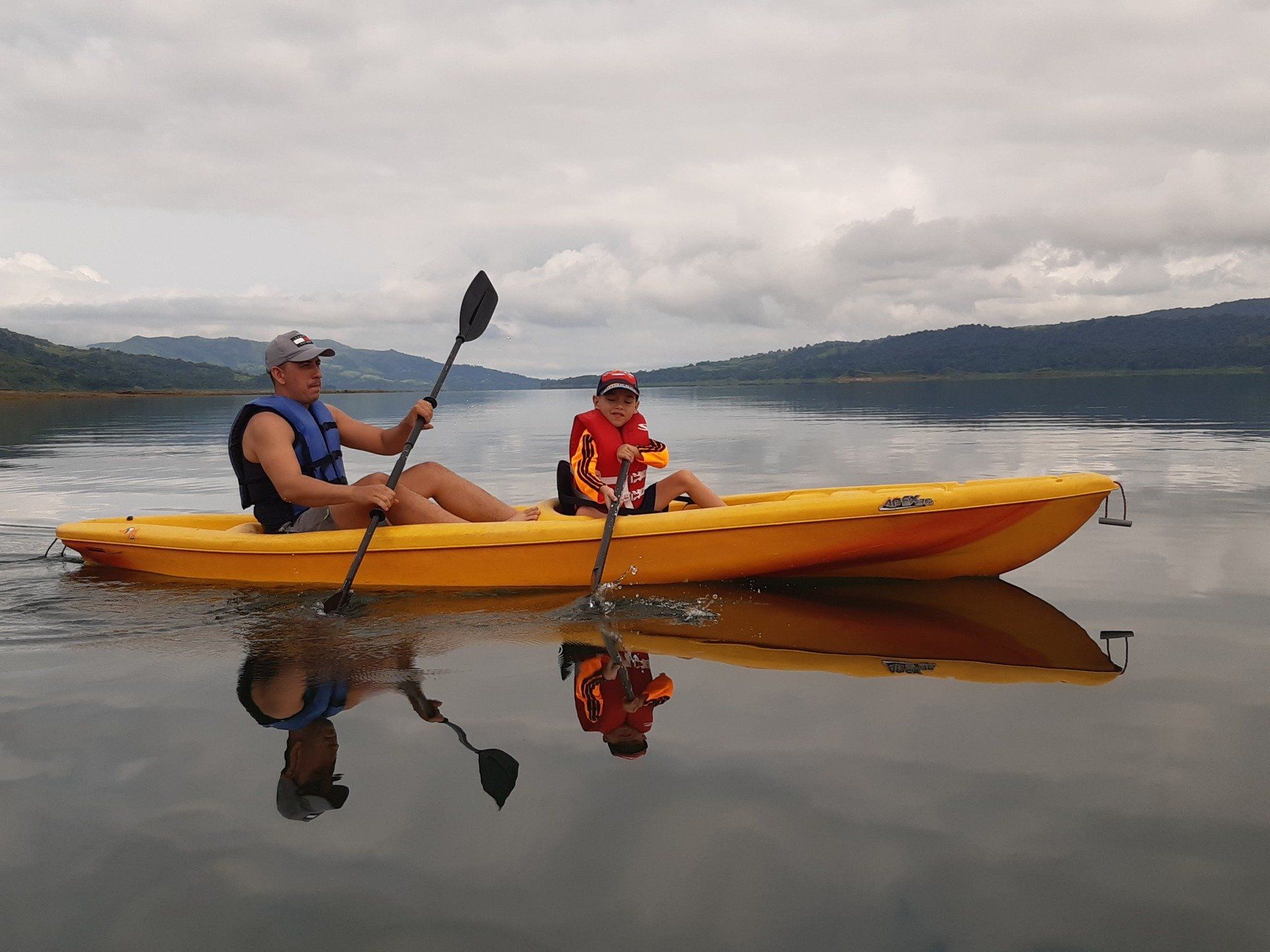 Father and child kayaking on calm water, under cloudy sky. Yellow kayak reflects in water.