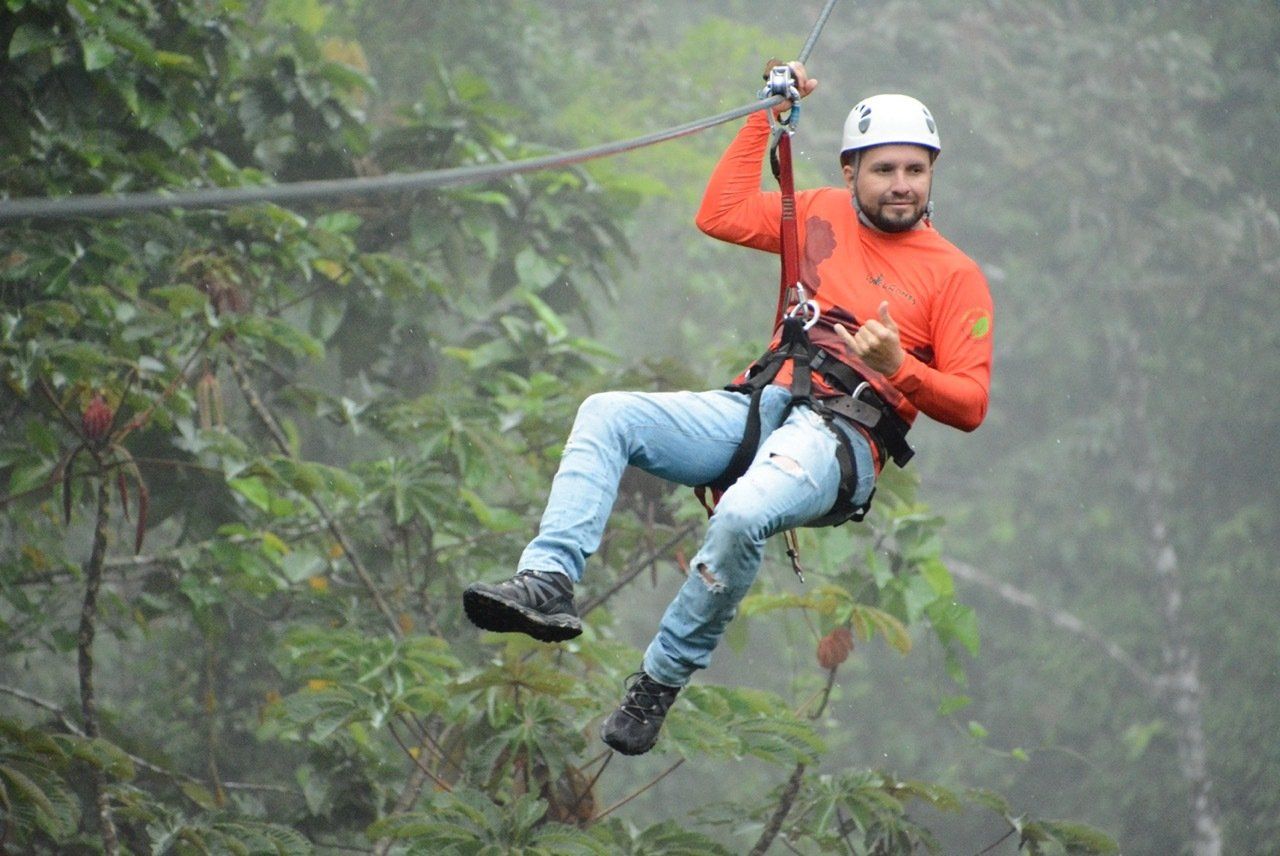 Man ziplining through a rainforest, wearing an orange shirt, jeans, and a helmet.