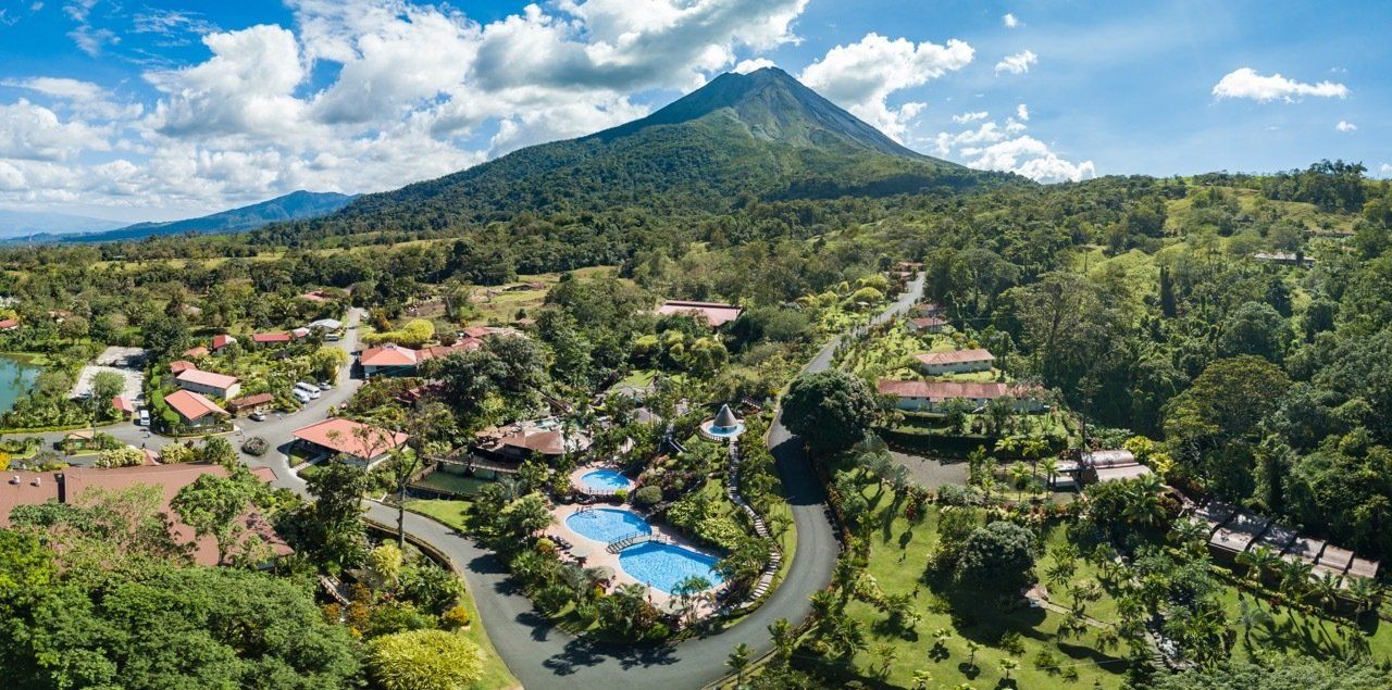 Aerial view of a resort with swimming pools and buildings at the base of a large, green, cone-shaped mountain.