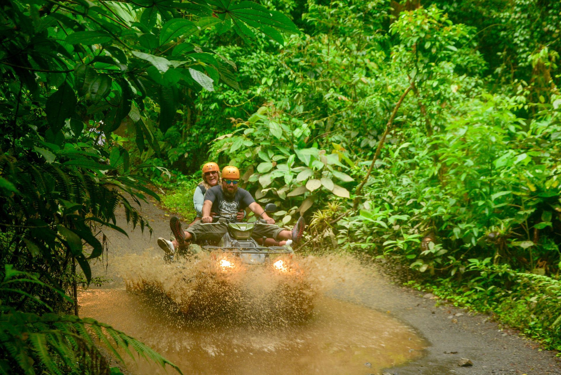 Two people on an ATV splash through a muddy puddle on a jungle road.