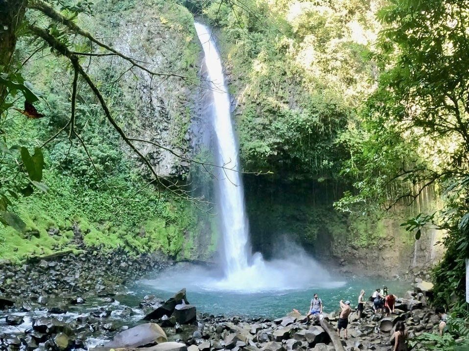 Waterfall cascading into a turquoise pool surrounded by lush green foliage; people at the base.