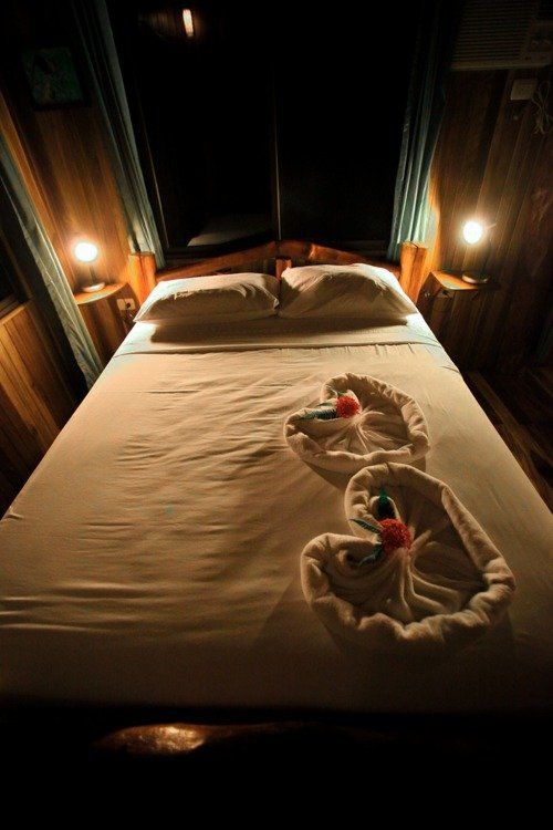 Bedroom with a made bed, folded towels, and lamps on each side. Wooden walls.