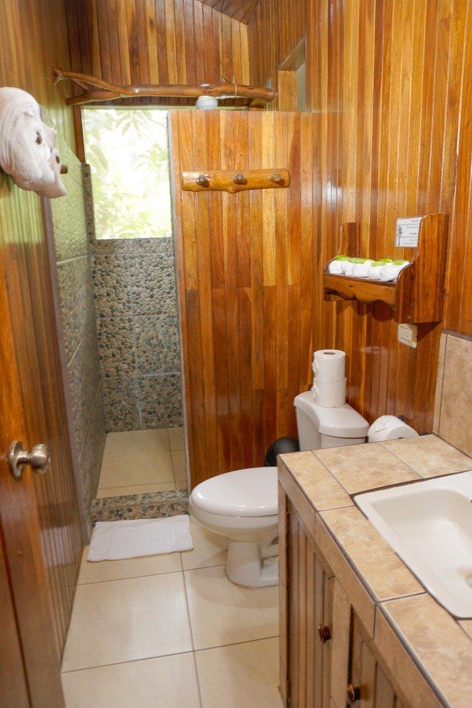 A wooden bathroom with a sink, toilet, and shower. Light shines through the window.
