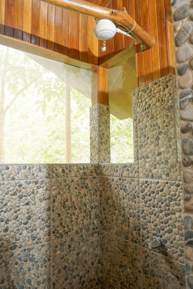 Shower with stone tile walls and wooden accents, looking out at green trees through a window.