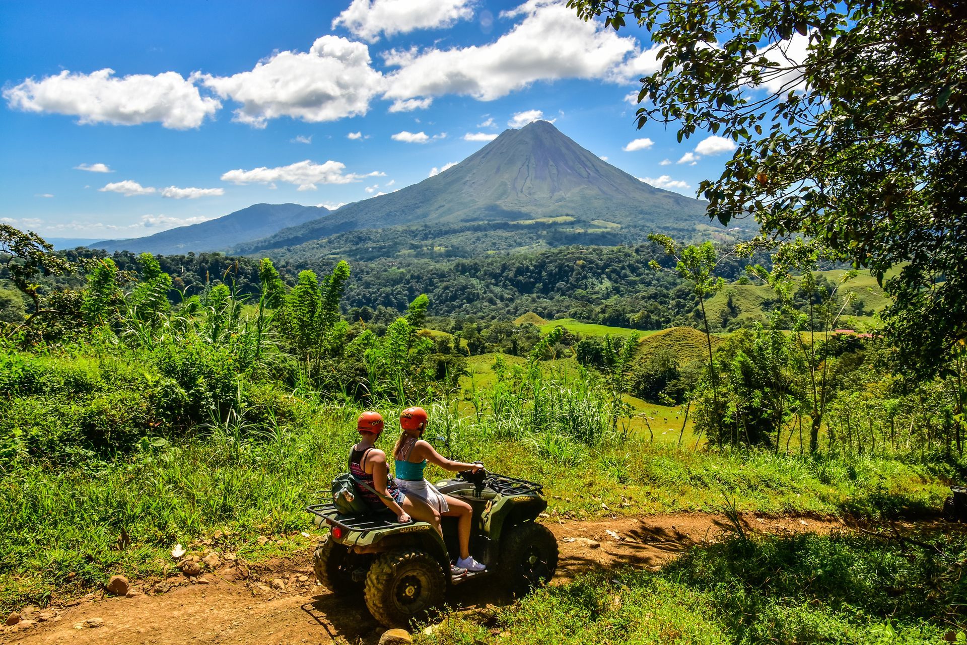 Two people riding an ATV on a dirt path, with a volcano in the background, Costa Rica.
