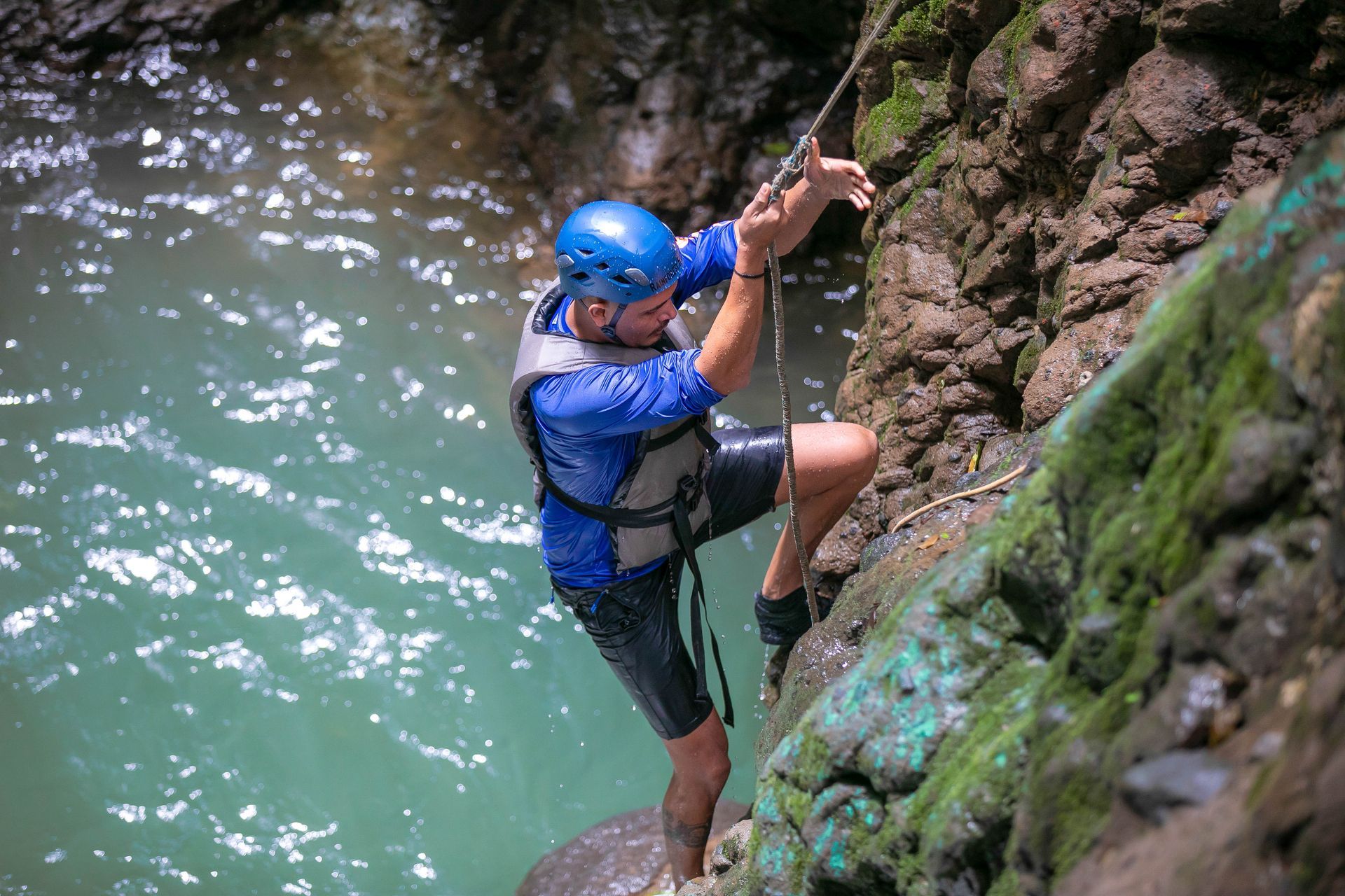 Man in blue climbing a rock face next to turquoise water, using a rope for assistance.